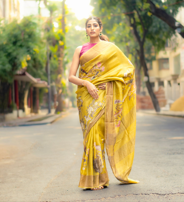 Woman in a yellow saree walking on a street with trees and buildings in the background
